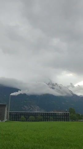 The video shows a landscape on a cloudy day featuring a large, flat-roofed building with many rectangular windows in front of a mountain range. The building is bordered by a green field. A chimney to the left of the building emits smoke or steam, and the mountain peak is partially obscured by low-hanging clouds. The sky is uniformly overcast. There are some trees on the right side of the building and on the mountain.