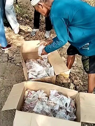 The video shows several people gathered outdoors, appearing to be in a wooded or unpaved area. Two large cardboard boxes filled with small, individually packaged items are visible. The items are mostly white or clear plastic bags, some containing what appear to be food items or possibly donations. One man, wearing a blue long-sleeved shirt and a white skullcap, is seen reaching into a box and handling the packages. Another person, wearing a white long-sleeved shirt, is also seen interacting with the boxes. The ground is covered with dirt and fallen leaves, and trees are visible in the background. The lighting suggests it is daytime.