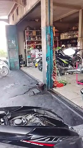 The video shows the interior of a motorcycle repair shop. The floor is made of gray tiles, and there are several motorcycles in various states of repair. In the foreground, a black scooter with its front panel removed is visible, revealing the engine components. To the left, a white motorcycle is parked. In the background, shelves are stocked with various parts and fluids, and a red air compressor hose is coiled on the floor. Multiple motorcycles, some with their engines exposed, are present throughout the shop. Banners advertising motorcycle brands and products hang on the walls. The lighting is dim and appears to be from fluorescent fixtures.