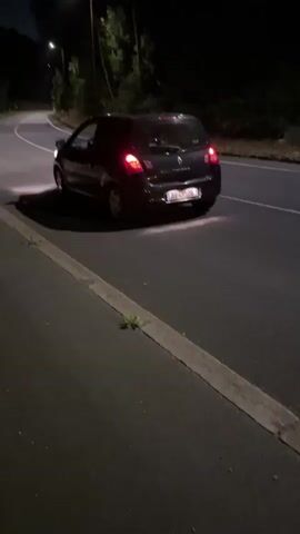 The video shows a dark colored car driving down a dark road at night, with streetlights visible in the distance. The car then pulls over and stops. A white car is seen with its trunk and driver's side door open. A spare tire is lying on the ground next to the white car. The car appears to be in the process of changing a flat tire.