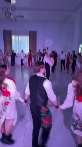 A group of people are dancing in a circle in a brightly lit room with a white floor and a marbled pattern. The room appears to be a banquet hall or reception area, with curtains in the background and a chandelier overhead. Most of the dancers are holding hands and moving in a circle, some are in traditional attire, others in formal wear. The camera is positioned low, focusing on the dancers' movements and the circular formation. Some individuals are more clearly visible than others due to motion blur. The overall atmosphere suggests a festive occasion, possibly a wedding or celebration.