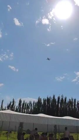 The video shows a bright, sunny day with a clear blue sky dotted with white clouds. The sun is prominent in the upper right corner of the frame. In the center of the sky, an airplane is visible in silhouette. Below the sky, a row of tall, dark green trees forms a line across the horizon. In front of the trees, there is a white, striped structure, possibly a canopy or awning, with some people visible at the bottom edge of the frame, their faces and upper bodies obscured. The airplane is in flight and appears to be the main subject of interest.