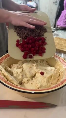 A person's hands are visible as they pour chopped chocolate and raspberries from a cutting board into a large bowl of cream and cake layers. The bowl has a red and yellow rim. A red knife is visible at the bottom of the frame. In the background, there is a layered cake on a white surface, a shelf with items, and a blurry figure in a purple coat. The scene appears to be a kitchen.