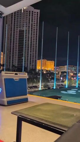 The video shows a night view of the Las Vegas skyline from a Topgolf location. The camera slowly pans right, revealing several tall buildings, including one with golden lights and the High Roller Ferris wheel. A blue Topgolf logo is visible in the lower right of the frame. Golf targets and lights are on the green below.