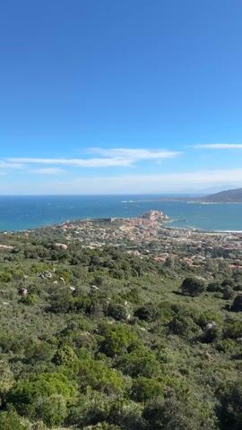 The video opens with a static shot of a scenic coastal landscape under a bright blue sky. In the foreground, there is lush green vegetation, consisting of trees and bushes, indicating a elevated viewpoint. Beyond the vegetation, a compact town with numerous buildings, many featuring red-tiled roofs, stretches along a peninsula that juts into a large body of deep blue water, likely the sea or an ocean. The water surface appears somewhat choppy, suggesting a breeze or wind. A distinct shoreline curves around the town and extends into the distance.

As the camera slowly pans to the right, more of the expansive bay is revealed. The town continues along the coast, and a wide sandy or rocky beach is visible where the land meets the water. Further in the background, a range of rugged mountains rises dramatically, their peaks appearing hazy in the distance. The sky above is a vibrant blue, adorned with scattered white, fluffy clouds that drift across the expanse. Sunlight illuminates the entire scene, creating clear visibility and highlighting the natural beauty of the area. The wind seems to be affecting the vegetation in the foreground, with some branches gently swaying. The video concludes on a wider view of the bay, the distant mountains, and the sky with its distinct cloud formations, with a prominent light green-leafed bush in the immediate foreground.