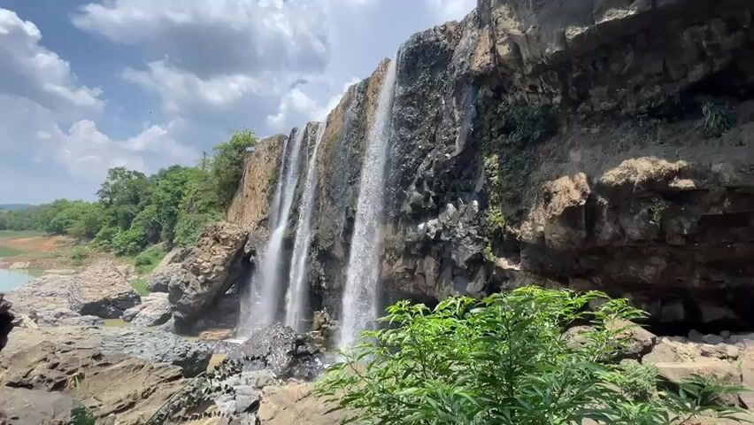 The video shows a waterfall cascading down a rocky cliff into a body of water. There are several streams of water falling, and the rocks are dark and wet. Green vegetation grows on the rocks and in the foreground. In the distance, a calm lake is visible surrounded by green trees. The sky is blue with white clouds. The video is shot from a low angle, emphasizing the height of the waterfall.