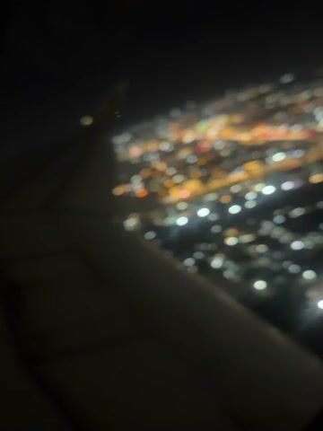 The video captures an aerial view of a city at night, seen from an airplane window, with the plane's wing visible in the foreground. The scene depicts the city of Addis Ababa, Ethiopia. Bright, sparkling city lights spread across the landscape below, illuminating the urban sprawl. Various colors of lights, including white, yellow, red, and green, dot the cityscape. The airplane wing, with its flaps and structures clearly visible, occupies the left side of the frame, indicating the plane is either descending for landing or ascending after takeoff. The night sky is dark, suggesting clear weather conditions. The camera slightly shifts and focuses, bringing the city lights into sharper detail after an initial blur.