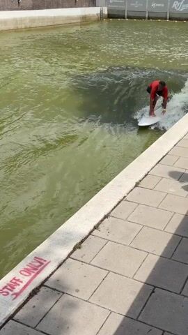 This media file captures a moment of surfing on a river wave in Rotterdam, Netherlands. The primary subject is a person on a surfboard, wearing a red shirt and dark shorts, actively riding a powerful wave. The surfer's body is positioned for balance and control, with arms extended to manage the ride. The water is a greenish hue, indicating it is likely a river or canal, and it's churning with white foam from the wave.

The scene is set alongside a paved walkway bordering the river. A concrete barrier runs along the edge of the walkway, and on this barrier, red spray paint spells out "START ONLY!". This suggests a designated area or a warning related to the surfing activity. In the background, a concrete wall forms the far bank of the river. The lighting suggests it is daytime, likely sunny due to the clear visibility and shadows. The overall atmosphere appears to be one of dynamic action and urban water sports. Another person's legs are visible in the water at the bottom of the frame, possibly waiting to surf or observing the activity.