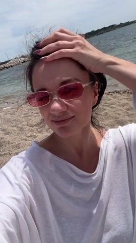 A young woman stands on a beach, facing the camera. She is wearing a white t-shirt and pink sunglasses. Her hair is pulled back. She touches her hair with her right hand. The beach has light brown sand, and the water is visible behind her. The sky is overcast.