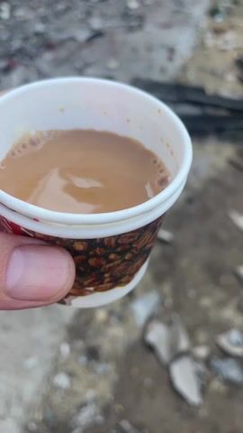 A hand holds a small paper cup of tea. The tea is light brown with small bubbles on the surface. The cup has a white rim and a brown pattern around the outside. The background is a cluttered courtyard with broken debris strewn about, including pieces of wood and stone. A brick wall surrounds the courtyard, and a building with barred windows is visible. The overall scene suggests a neglected or abandoned space.