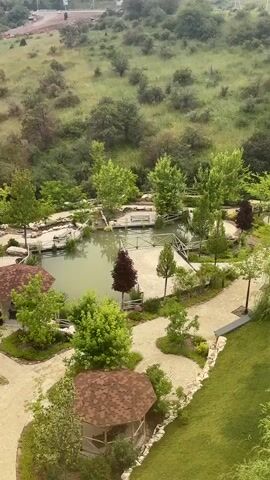 This video shows a drone shot from above a park-like area with a pond, gazebos, and pathways. The park is surrounded by rolling hills covered in greenery and trees. In the background, a road winds through the hills, with some construction activity visible in the distance. The weather appears overcast.
