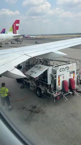This is a video taken from inside an airplane looking out the window at an airport tarmac. A refueling truck, branded "carboil," is positioned under the wing of the airplane. A worker wearing a yellow vest and dark pants is standing near the truck, possibly overseeing the refueling process. The truck is connected to the airplane, and a hose is visible. In the background, other airplanes are parked, including one with the "Air Portugal" logo on its tail. The sky is partly cloudy. The video shows the truck fueling the airplane.