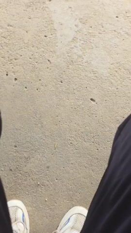 The video shows a concrete surface, likely a patio or sidewalk, with cracks and minor debris. A wooden post with a concrete base is visible, supporting an unseen structure above. In the background, there is a lawn, a low concrete wall, small shrubs, and a blurred view of houses across the street. The video starts with a view of a person's leg in dark clothing, then pans upward to show the scene. The camera is handheld, and the movement is slightly shaky. There is no other activity or interaction visible.