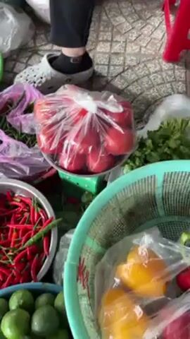 The video shows a collection of vegetables and other food items at what appears to be an outdoor market.  A person wearing a dark tracksuit, grey shoes, and socks is visible in the lower left corner. A second person, wearing a red garment, is also present but mostly obscured from view.  A hand, presumably belonging to one of the individuals, reaches into a light green basket to retrieve a plastic bag containing yellow bell peppers.  The person then closes the bag with a knot.  Other items include tomatoes, cilantro, chili peppers, limes, onions, red onions, bananas, garlic, and ginger, all arranged in baskets and plastic bags.  The ground is tiled with a grey geometric pattern.