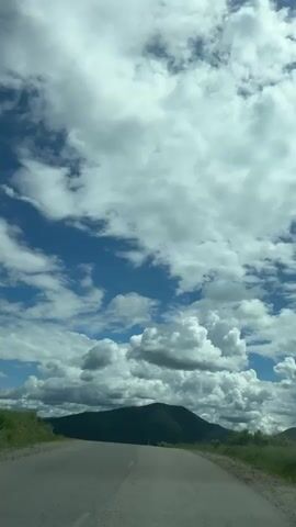 A video shows a road with a mountain in the distance under a cloudy sky. The road appears to be asphalt with a white line marking the center. The sky is blue with scattered white clouds. The mountain is covered in green vegetation. The camera is mounted in a vehicle, as indicated by the presence of the vehicle's hood in the foreground. The video is shot from a first-person perspective, looking forward along the road. The time stamps indicate a short video, approximately 4 seconds long.