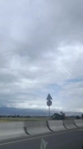 A view from inside a vehicle on a cloudy day. The road is visible in the foreground, with a concrete barrier to the left. Beyond the road, there is a grassy field and some trees. In the distance, mountains are visible under a cloudy sky. Several contrails from airplanes are streaking across the sky.