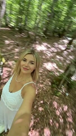 A blonde woman wearing a white dress and a pearl necklace is walking through a forest. She is holding a phone in her right hand, appearing to film herself as she walks. The camera is moving with her, capturing her from the chest up. The background is a blur of green leaves and trees, indicating movement and a wooded environment.