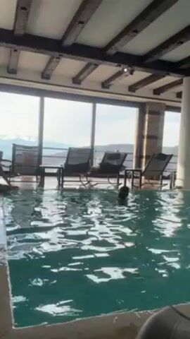 The video shows a person swimming in an indoor pool that overlooks a mountain range. The pool is surrounded by lounge chairs and a large window. The person in the pool is moving around. The mountains are visible through the window, with some snow-covered peaks. The ceiling of the room has wooden beams.