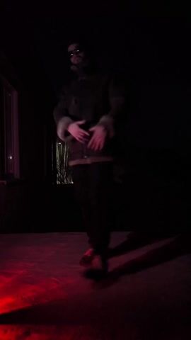 The video shows a person dancing in a dimly lit environment with a red light illuminating the ground. The person is wearing a dark jacket with a light-colored lining, dark pants, and black shoes with red laces and soles. They are also wearing dark sunglasses and a hat. The person is moving their arms and legs in a rhythmic manner, seemingly enjoying the music. Behind them is a dark building with a window. The scene is dark, with only the red light providing illumination to the dancing figure and the ground around them.