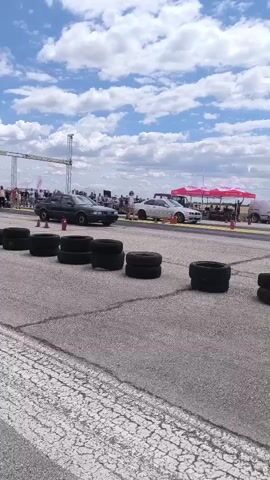 The video shows a drag race. Two cars, a dark colored pickup truck and a lighter colored sedan, are lined up at the start of a drag strip. The track is asphalt with painted white lines. Several stacks of tires are placed along the sides of the track, acting as markers or safety barriers. Spectators are visible in the background, standing behind a barrier. Tents and structures, likely for the event, are also in the background. The sky is blue with scattered white clouds. The cars then accelerate down the track. The pickup truck pulls ahead. The camera pans to follow the cars as they race. Smoke is visible coming from the tires of the pickup truck as it launches. The sedan also launches, but appears to be slower. The video ends as the cars are moving down the track.