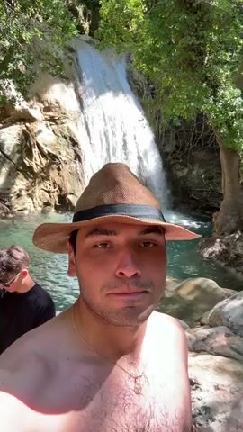 A man in a straw fedora with a dark band is taking a selfie in front of a waterfall. He is shirtless, wearing a gold necklace with a pendant. Behind him, another person, partially visible, is standing in the clear, turquoise water. The waterfall cascades down rocky terrain covered in lush green foliage. The scene appears to be in a natural, outdoor setting, likely a park or nature reserve, given the presence of the waterfall and abundant plant life. The lighting suggests it is daytime, with sunlight filtering through the trees. The man's expression is neutral, but the setting evokes a sense of leisure and enjoyment of nature. The overall impression is a vacation or recreational outing in a beautiful natural landscape. There is no visible text in the media file.