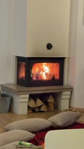 A static, full shot shows a fireplace burning in a corner of a room. The fireplace is white with a dark metal framed glass front. Logs are stacked beneath the firebox. In front of the fireplace on the wooden floor is a red and black plaid blanket with several square beige cushions. Next to the blanket is a small white table with items on it. To the left of the fireplace is a gray bucket. Sounds of children talking can be heard.