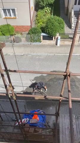 A person is using a hose to spray water on the ground from a scaffolding. They are wearing a dark shirt and shorts. The ground is wet and there is a shadow on the ground cast by the person and the scaffolding. There are houses and trees in the background.