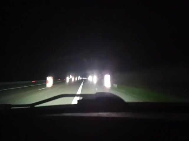 The video shows a car driving on a highway at night. There are lane dividers and construction signs, with lights visible ahead. The view is from inside the car, through the windshield.