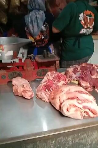 A close-up, eye-level shot of a butcher's counter displays several large cuts of raw meat. In the foreground, a large, marbled cut of beef with prominent white fat is positioned on the right. To its left, a smaller, more lean cut of red meat is visible. Further to the left, a pale, less distinctly veined cut of meat rests near a red, antique-style balance scale with a silver weighing pan. Behind the meat, a person in a green shirt and apron is partially visible, seemingly engaged in work. Another person, wearing a headscarf with a blue and grey pattern featuring an orange and black graphic, is also in the background. The environment appears to be a market stall, with what looks like bags of food items hanging from the ceiling in the upper background. The lighting is dim, creating a somewhat dark and shadowed atmosphere.