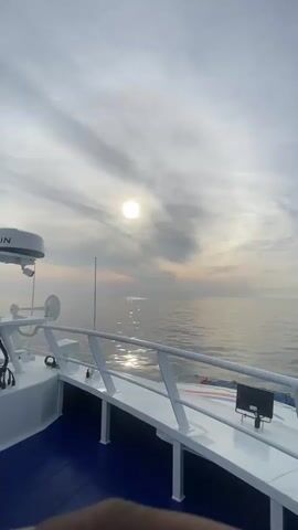 The video shows a view from a boat deck looking out over the ocean. The deck is white with blue flooring and has white railings. A radar dome is visible on the left side of the frame. The sun is visible through a cloudy sky and is reflected in the water. The water is calm and there is a slight haze on the horizon.