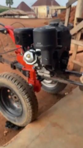 The video shows a close-up view of a red and black walking tractor parked on a dirt surface. The walking tractor has two large wheels with treads and a visible engine. In the background, there are some buildings with red roofs, trees, and a person riding a motorcycle. The environment appears to be rural or semi-rural. There are no people interacting with the walking tractor in the foreground. The lighting suggests it is daytime, possibly overcast.