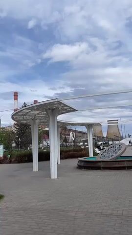 The video presents a slow, horizontal pan across a modern public park or square in Urussu, Russia, under a cloudy sky. The ground is paved with interlocking bricks.

Prominently featured are several contemporary white architectural structures resembling pergolas or canopies, supported by thick white pillars. Their roofs are flat and circular or semi-circular, adorned with intricate, laser-cut patterns. In the foreground, a circular stone-like structure, possibly a decorative fountain or planter, sits on the paved area, topped with a large, ornate metal lid featuring geometric cutouts.

In the mid-ground, a children's playground area is visible, equipped with colorful swings and other play structures. Several people can be seen in the park: one person walks near the playground, another is on a swing, and others stroll further in the distance. Benches are scattered along the paved pathways, which are bordered by green spaces with bushes and leafless trees, suggesting early spring or late autumn.

In the background, two large, grey cooling towers characteristic of a power plant dominate the skyline, hinting at the industrial context of the location. The overall scene depicts a clean, well-maintained public space designed for leisure and recreation, juxtaposed with an industrial backdrop. There is no discernible text visible in the video.