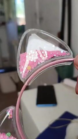 A close-up shot focuses on a pink headband with clear plastic cat ears filled with pink glitter and small star-shaped sequins. A hand is visible on the right side of the frame, holding one of the ears. The background is blurred, showing a room with a white table, a blue smartphone, and some objects on shelves. The lighting appears to be natural, coming from the left side.