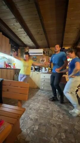Inside a cozy cabin with wooden beams and paneling, three people are dancing to upbeat music. A man in a blue shirt and two women, one in a yellow shirt and the other in a blue top, are linked arm-in-arm. They are moving in a circle with spirited enthusiasm. The cabin features a kitchen area, complete with cabinets and appliances, and a faux fireplace on the television screen. A wooden bench sits nearby, and the floor is tiled. The overall atmosphere is lively and warm.