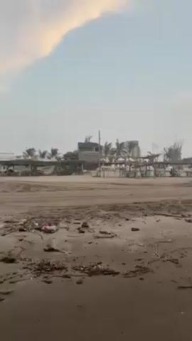 The video shows a beach on what appears to be a hazy day, with a gray sky tinged with pink. The sand is dark and damp, littered with debris including plastic trash, wood, and rope. Structures with thatched roofs and green chairs are lined up along the beach. Several cars, including what appears to be a Tesla, drive along the beach. In the distance, a few individuals are walking near the water's edge. The camera pans across the scene, revealing more of the shoreline.