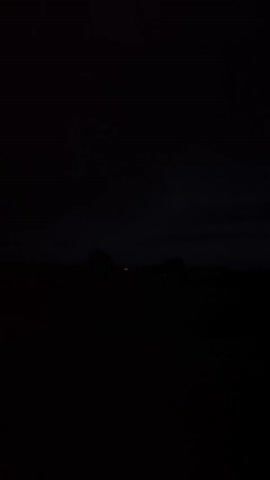 The video is extremely dark and grainy, making it difficult to discern any clear details. The scene appears to be an outdoor environment at night. There are faint lights visible in the distance, possibly from buildings or streetlights, suggesting a rural or suburban area. The sky is dark and cloudy, and there is a brief flash of light, possibly lightning, in the distance. The foreground is completely dark, obscuring any ground features or objects. The video quality is poor, with significant noise and low resolution.