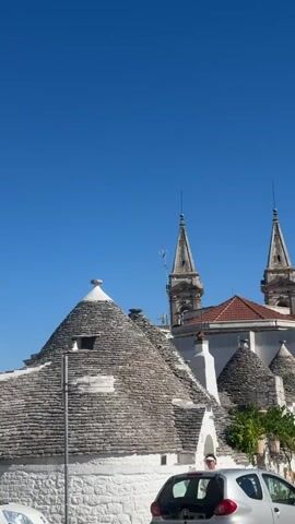 A drone-like shot pans across a town street with distinctive cone-shaped roofs, characteristic of the Puglia region of Italy. In the foreground, a small, silver car is parked on the left side of the street. Further down the street, several people are visible walking. In the background, two church steeples rise above the buildings against a clear blue sky. The architecture features whitewashed walls and conical stone roofs, with a few trees and shrubbery interspersed. The road appears to be paved with cobblestones. The overall impression is a sunny day in a historic European town.