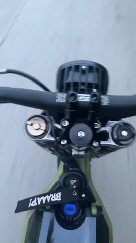 The video is a first-person perspective shot from a moving vehicle, likely a motorcycle or dirt bike, as indicated by the handlebars and front suspension visible. The vehicle is traveling on a paved road with some cracks and rough patches. The camera is pointed forward and slightly downwards, showing the road ahead, the handlebars, and the front of the vehicle, including a black headlight. The background consists of a blurry gray road and a hint of green foliage on the right side. The footage appears to be taken during the day. There are no other people or vehicles visible. The overall impression is of riding along a somewhat rural or less maintained road.