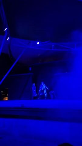 A group of people in costumes perform on a stage. The stage is lit with alternating blue and red lights, with smoke present in the background. The performers are engaged in what appears to be a choreographed dance or dramatic scene. The stage is an outdoor setting, possibly an amphitheater, with a white roof structure overhead.