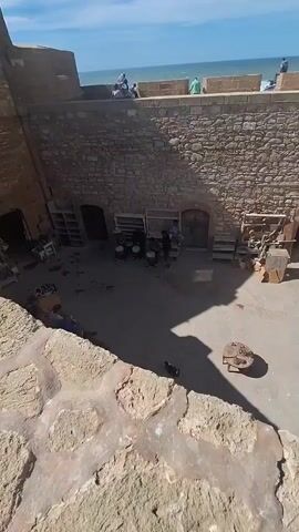 The video shows a courtyard within ancient stone walls. In the foreground, a textured stone wall occupies the bottom portion of the frame. Beyond this, the courtyard is visible, paved with stone. On the left side of the courtyard, there are shelves with various items, possibly merchandise. In the center, a group of people are gathered around a collection of drums, some of whom appear to be playing them. To the right of the drums, there are more shelves and some wooden structures. Archways are visible in the back wall, leading to darker interior spaces. Above the courtyard, the sky is blue with scattered clouds, and the sea is visible in the distance, with people standing on the parapet of the outer wall overlooking the water. The overall impression is of a historic outdoor space, possibly a market or performance area, with a view of the ocean.