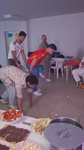 In an indoor setting, a group of men engages in a playful activity amidst a buffet setup. Three men are positioned on the floor, with the man at the back supporting the one in front of him, forming a kind of "human chain" doing push-ups. They are attempting to pick up objects from the floor with their mouths. Observers stand and sit around, one of whom is clapping his hands in encouragement. The buffet table in the foreground contains an array of meats and cheeses. The flooring is tiled, and the walls are painted a neutral color. There's a relaxed and jovial atmosphere in the room.