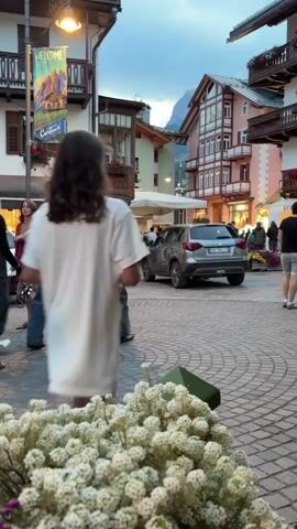 The video captures a bustling street scene in a European town square, likely in the Alps, given the architectural style of the buildings and the mountainous backdrop. The time of day appears to be late afternoon or early evening, with the sky a soft blue and illuminated by streetlights and building lights.

In the foreground, a large cluster of white, fluffy flowers, possibly hydrangeas or astilbe, fills the bottom of the frame. The cobblestone pavement is visible throughout the scene, marked with circular patterns.

A group of people are walking through the square. In the center, a woman in a white t-shirt dress and a man in a white t-shirt with graphics on the back are walking away from the camera. They appear to be part of a larger group of young adults. Another woman in a white shirt and blue jeans is walking towards the camera, her back partially to the viewer. Several other individuals, dressed in casual attire, are scattered throughout the square, some walking, some standing.

The buildings lining the square exhibit a distinctive Alpine architectural style, with multi-story structures featuring pitched roofs, balconies, and wooden accents. Some buildings have storefronts at ground level, with illuminated windows displaying goods. A banner with the word "WELCOME" and images of mountains hangs from a lamppost on the left side of the frame.

A silver car is parked on the right side of the square, and another white car is partially visible on the left. The overall atmosphere is one of a lively, pedestrian-friendly urban environment.