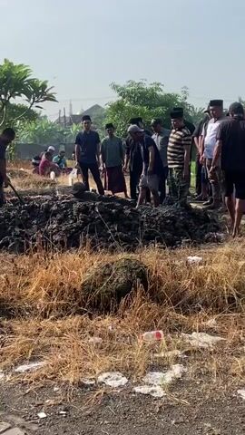 The video shows a group of around 15 men standing around a mound of black soil in a field of dry grass on a sunny day. Several of the men are digging into the soil with shovels. Other men watch and are dressed in a variety of clothing, including traditional Muslim attire and casual wear. In the background, there is a building with a golden dome, possibly a mosque, as well as trees and power lines. The overall scene appears to be a burial or excavation taking place in a rural or semi-rural area.