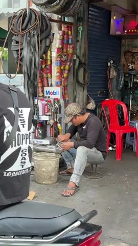 A man in a camouflage baseball cap, black long-sleeved shirt, light blue jeans, and flip-flops sits on a stool in front of a shop. He is working on a metal object with his hands, holding it over a grey bucket. The shop is filled with shelves stacked with various items, including tires, cans of oil, and other automotive parts. To the left, a portion of a motorcycle and a person wearing a black t-shirt with white lettering are visible. To the right, a bright red plastic chair and several small blue plastic stools are placed outside the shop entrance, along with a motorcycle in the background. The ground is paved with concrete and appears wet in places.