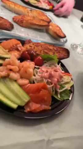 A video shows a group of people at a dining table with plates of food and drinks. The table is covered with a light green tablecloth. Each plate contains avocado, cucumber, shrimp, salmon, salad, and what looks like toasted bread.  A white bowl with a pink sauce and an orange spoon rests on the table. There is a wine bottle, wine glasses, and a small stack of red napkins also on the table. People around the table are seen eating and talking.