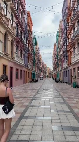 A person walks down a street lined with colorful buildings. The person is wearing a brown top and a white skirt. The street is paved with tiles. There are scooters parked along the sides of the street. There are string lights hanging across the street. The sky is cloudy.