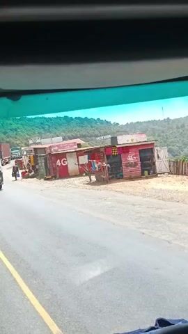 A person is driving a truck on a paved road, indicated by a yellow line, passing small roadside businesses. There are multiple trucks ahead, some with tarpaulin covers, and one is a white Mercedes-Benz with the license plate KCW 008P. The businesses are small structures, some painted red with "4G" displayed on them. The landscape includes green hills in the background. The video is shot from inside the truck, capturing the windshield and a portion of the dashboard, including a blue cloth. The camera shakes slightly, indicating movement. The driver's hands are visible on the steering wheel.