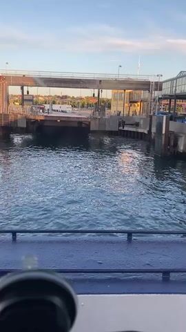 A video captures a body of water with choppy waves. In the background, a ferry terminal is visible. The terminal features a structure spanning across the water, likely a bridge or ramp for vehicles. There are buildings on the right side of the terminal, and some lights are visible on the structure. The sky above is a soft blue with some scattered clouds, and the overall lighting suggests it might be late afternoon or early evening. The foreground shows the edge of a blue surface, possibly the deck of a boat, with a dark circular object in the bottom left corner.