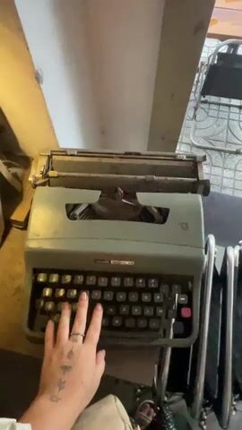 A close-up, overhead shot shows a person's left hand with a ring on their ring finger and tattoos on their arm typing on a vintage Olivetti Lettera 32 typewriter. The typewriter is a sage green color with black keys and a red carriage return lever. The person's fingers are pressing down on the keys. The typewriter is on a dark surface, possibly a table or desk. In the background, out of focus, are parts of chairs and a wall. The lighting is dim.