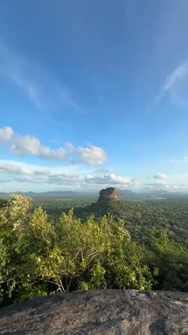 The video shows a panoramic view of a lush, green landscape under a blue sky dotted with white clouds. In the distance, a large, dark rock formation rises from the forest. The foreground consists of vegetation and a large rock formation from which the view is being captured. The overall impression is one of natural beauty and tranquility.